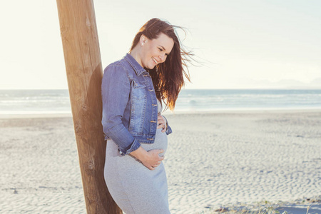 快乐怀孕的年轻母亲站在户外的一根杆子上，俯视和欢笑的海滩场景，带着休闲装的侧面景色。