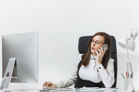 美丽的严肃的商务妇女在西装和眼镜坐在办公桌上, 在现代计算机与文件在轻型办公室, 谈论手机解决问题, 在白色背景