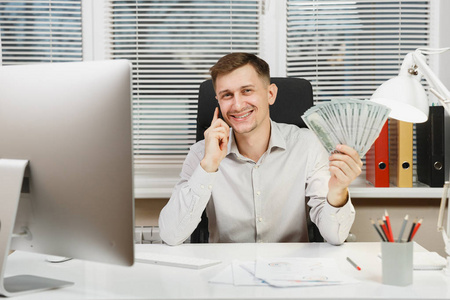 电脑赚钱英俊微笑的商人穿着衬衫坐在办公桌上, 用大量的现金赚钱, 在