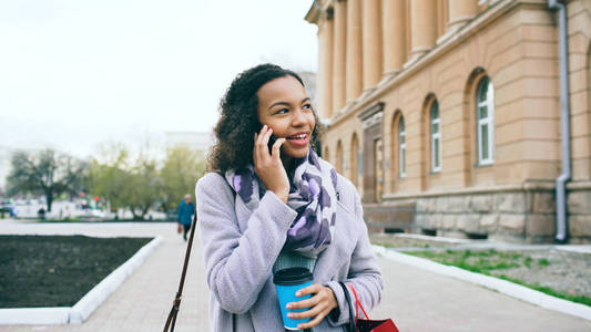 有吸引力的混合种族女孩谈论智能手机和喝咖啡散步在城市街道与袋子。购物的少妇在购物中心销售