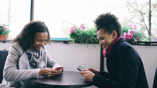 两个具有吸引力的混合的种族女性朋友一起在街边咖啡馆的户外使用智能手机共享