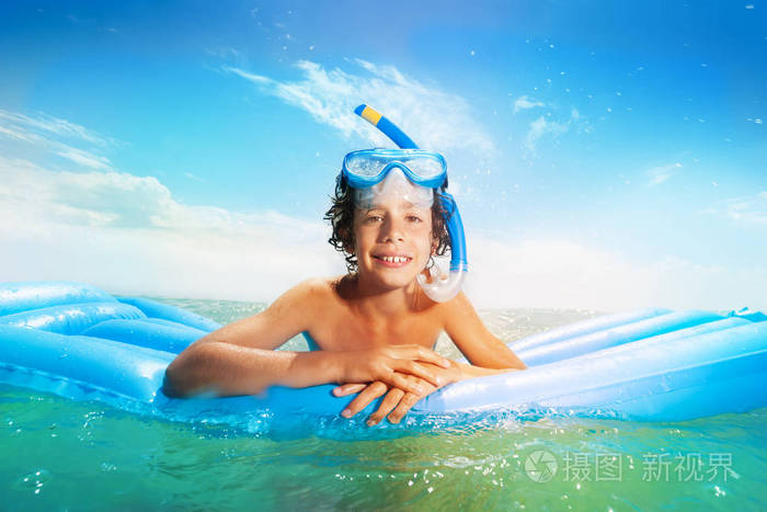 戴着潜水面罩的好男孩在海中的蓝色充气垫子上游泳