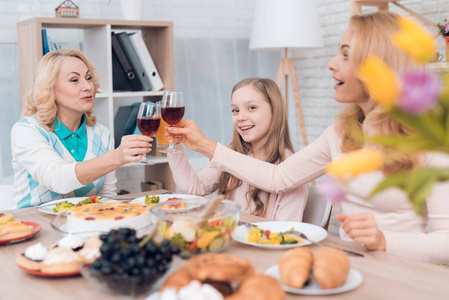 妈妈和奶奶在喝葡萄酒，一个小女孩在喝果汁。 他们在家吃晚饭。 他们桌子上有蔬菜和糖果。