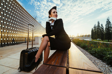 时尚美丽的年轻女子空姐穿着制服坐在长凳上, 等待她的飞行, 与手提箱