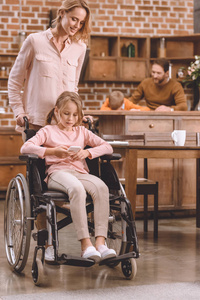 微笑的母亲看着残疾的女儿坐在轮椅上，在家里使用智能手机