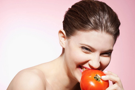 微笑着美丽的白种人年轻女人在粉红色的背景下吃番茄