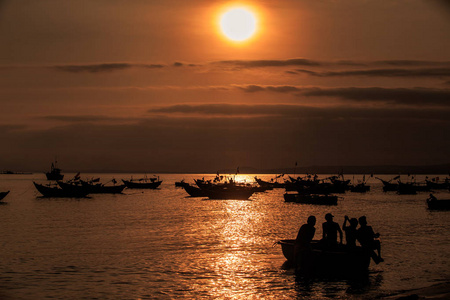 在宁静的海洋海湾上方的夜空中，巨大的橙色太阳盘在云层中，船和人在日落时的轮廓