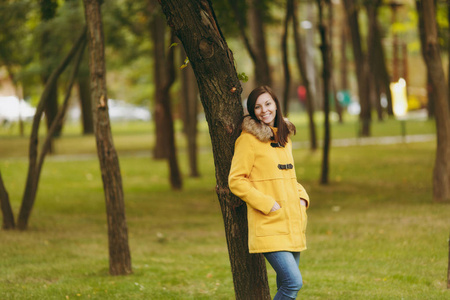美丽快乐白种人年轻微笑的棕色头发的妇女在黄色外套, 牛仔裤, 靴子在绿色森林。秋金叶时尚女模特在初秋公园户外站立行走