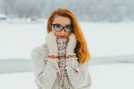 在下雪时戴着眼镜手套和围巾的迷人红头女郎的肖像。草甸地点
