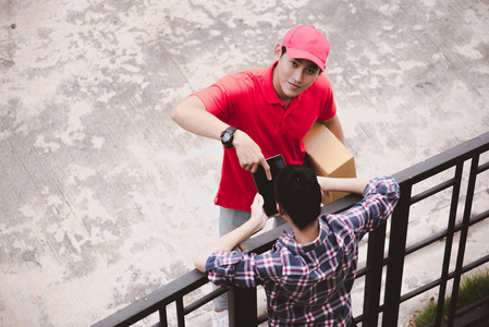 年轻的后勤职业概念。 快乐送货员把他的包裹给家里的顾客。 在真正的房子里。 亚洲中国人适合穿红色马球衫和牛仔裤，二十出头戴红色帽