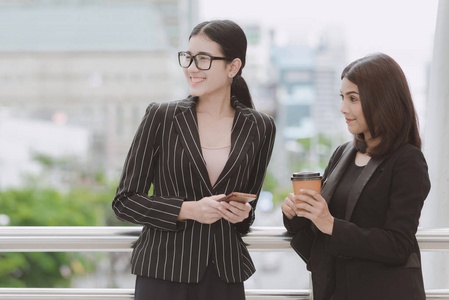 两位美丽自信的亚洲商界女性在休息时间喝杯咖啡。 两名穿办公室西装的妇女在办公室休息时间交谈，在户外自然光线下进行。 职业女性概念