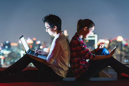 年轻的亚洲夫妇使用笔记本电脑和智能手机,彼此背靠背