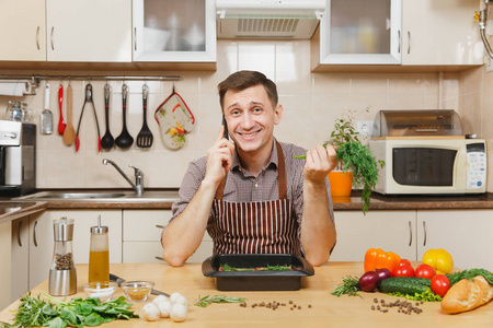 穿着围裙的人坐在餐桌旁, 吃着蔬菜, 莳萝, 用手机聊天, 在家做饭, 用