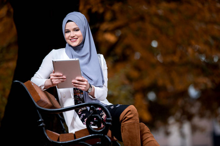 美丽的年轻女孩戴着头巾，用平板电脑在公园长凳上放着秋色的东西