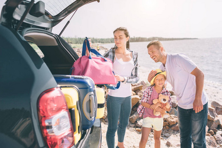 一对年轻的夫妇站在开着的汽车的行李箱和行李箱。爸爸妈妈和女儿在海边或海边或河边旅行。汽车夏季乘车