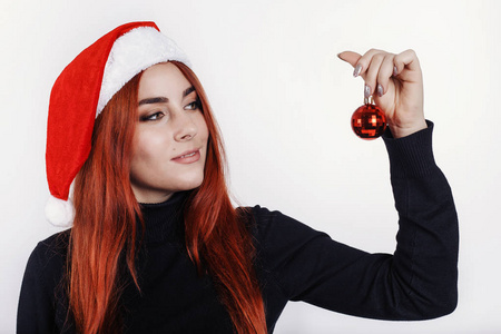 戴着圣诞帽的年轻红发女郎手里拿着一个小小的圣诞球，看着它