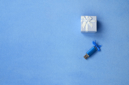 明亮的蓝色USB记忆卡与蓝色蝴蝶结躺在一个蓝色的小礼品盒旁边，一个小蝴蝶结在柔软和毛茸茸的浅蓝色羊毛织物的毯子上。 经典女性礼品