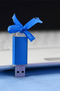 明亮的蓝色USB闪存卡与一个蓝色蝴蝶结躺在一条柔软和毛茸茸的浅蓝色羊毛织物毛毯旁边的白色笔记本电脑。 经典女性礼品设计的记忆卡