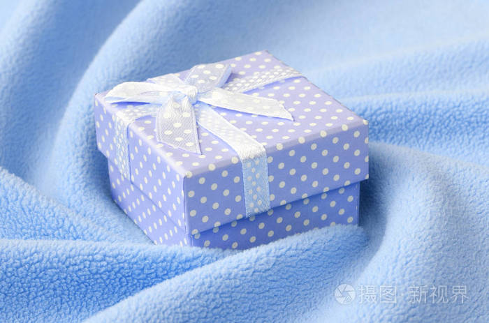 一个蓝色的小礼品盒，有一个小蝴蝶结，躺在一条柔软而毛茸茸的浅蓝色羊毛织物的毯子上，上面有很多浮雕褶皱。 打包礼物送给你可爱的女朋