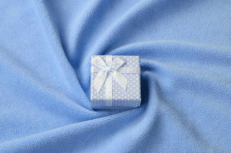 一个蓝色的小礼品盒，有一个小蝴蝶结，躺在一条柔软而毛茸茸的浅蓝色羊毛织物的毯子上，上面有很多浮雕褶皱。 打包礼物送给你可爱的女朋