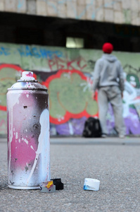 使用过的气溶胶喷漆罐，上面有粉红色和白色的油漆，躺在沥青上，对着涂鸦画中的墙前站立的人。