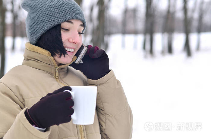 冬天公园里漂亮迷人的高加索穿洞女孩的肖像。 她正在谈论现代智能手机和喝咖啡。 青年街头时尚。 在露天散步