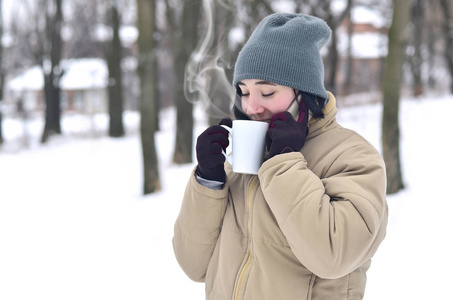 漂亮的年轻女孩戴着温暖的冬帽和外套。 有趣的黑发女性模特在寒冷的天气里带着智能手机和咖啡杯走在外面。 微笑快乐的模特摆姿势。 好