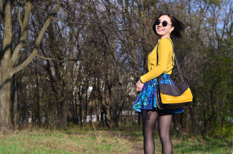 漂亮可爱的黑发女孩穿着一件亮黄色的毛衣，带着一个黑色的袋子，戴着圆形的黑色太阳镜，穿过春天的森林。 阳光明媚的春天，一个快乐的黑