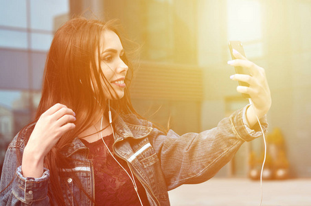 年轻的黑发女孩穿着牛仔裤，在一栋办公楼的背景下，在智能手机上自拍。在手机摄像头上拍摄自己的时尚概念
