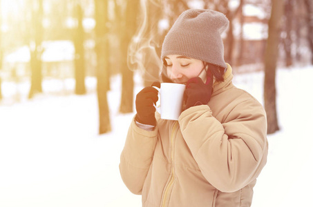 漂亮的年轻女孩戴着温暖的冬帽和外套。在寒冷的天气里，有趣的黑发女性模特带着智能手机和咖啡杯走在外面。微笑快乐模特摆姿势。好天气