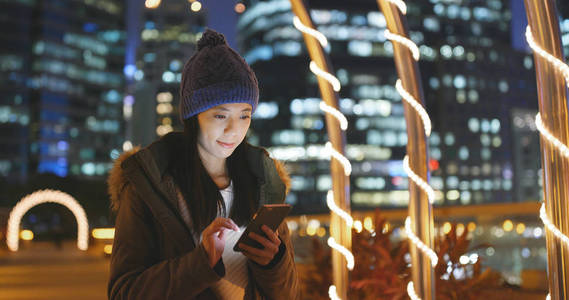 女人晚上在城市里使用智能手机，在城市背景的智能手机上上网冲浪，女人穿着冬季夹克