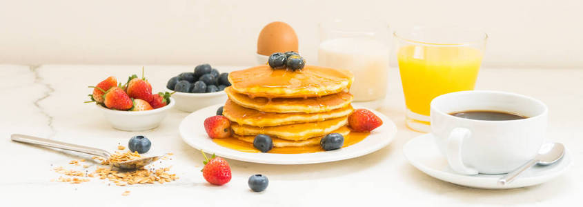 健康早餐套餐，包括煎饼和麦片蓝莓草莓和黑咖啡牛奶和橙汁，背景白石桌