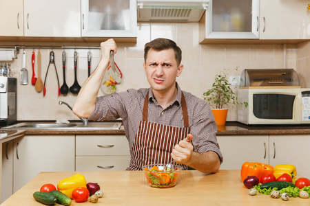 愤怒的疯狂饥饿的年轻人在围裙, 棕色的衬衫坐在餐桌上, 用叉子和刀子在光厨房攻击蔬菜沙拉。节食概念。健康的生活方式。在家做饭。准