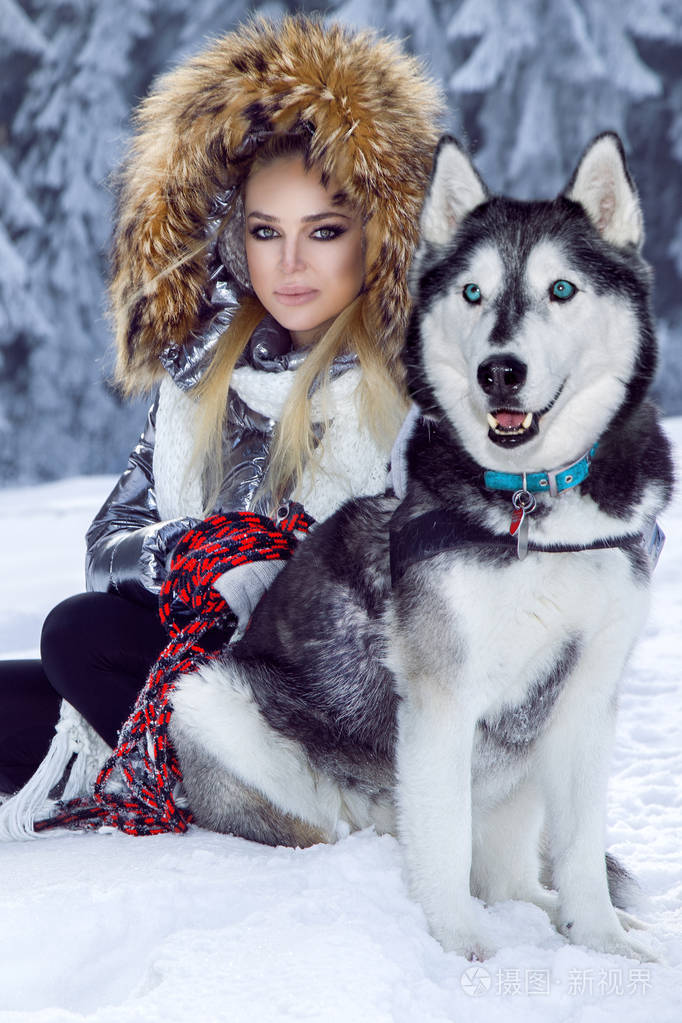 美丽的金发女人站在雪地上抱着哈士奇的狗背景是美丽的山和雪的景色