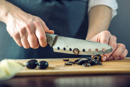 黑围裙的主厨用小刀切黑橄榄。环保型烹饪产品概念