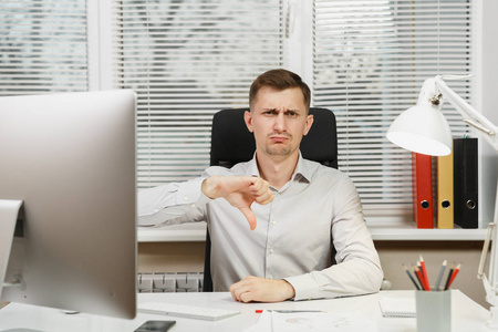 严重疲惫和压力的商人坐在办公桌前的衬衫, 显示拇指下来, 工作在计算机与现代显示器, 文件在轻型办公室的窗口背景。经理或工作人员