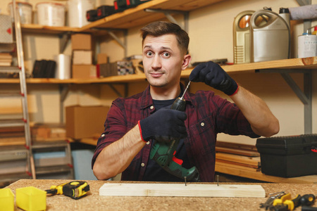英俊的微笑高加索青年男子在格子衬衫, 黑色 t恤, 手套钻与权力钻在一块木头, 工作在木工车间在木桌地方用不同的工具