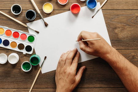 艺术家的手紧紧地拿着画笔在一张白纸上，靠近打开的彩色颜料罐和棕色木桌上的调色板，准备用水彩作画。 创造力的概念。
