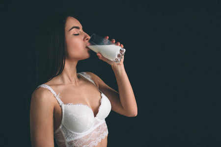 女性模型喝牛奶在白色背景。年轻妇女与长的黑发站立隔绝在黑背景。这个女孩有一个运动的身影, 她穿着白色的内衣