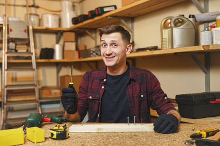 英俊的微笑高加索青年男子在格子衬衫, 黑色 t恤, 手套扭螺丝, 在木工车间工作在木制桌子的地方与一块木头, 不同的工具