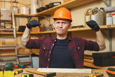 英俊强壮的高加索年轻人在格子衬衫, 黑色 t恤, 橙色防护头盔, 在木工车间工作的手套在木制桌子的地方与一块木头, 锤子, 不同