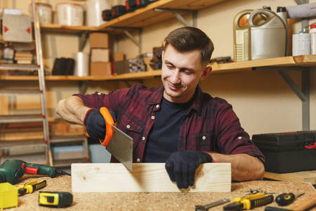 英俊迷人的微笑高加索青年男子在格子衬衫, 黑色 t恤, 手套锯木材与锯, 在木工车间工作在木桌地方用一块木头, 不同的工具