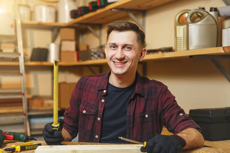 英俊的高加索青年男子在格子衬衫, 黑色 t恤, 手套测量木材的长度与卷尺, 在木工车间工作在木桌地方用不同的工作工具