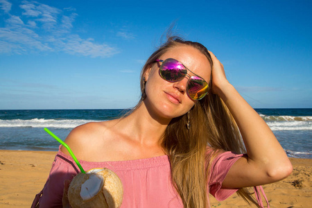 戴着墨镜的年轻女孩走在夏威夷考艾岛的海滩上喝椰子汁