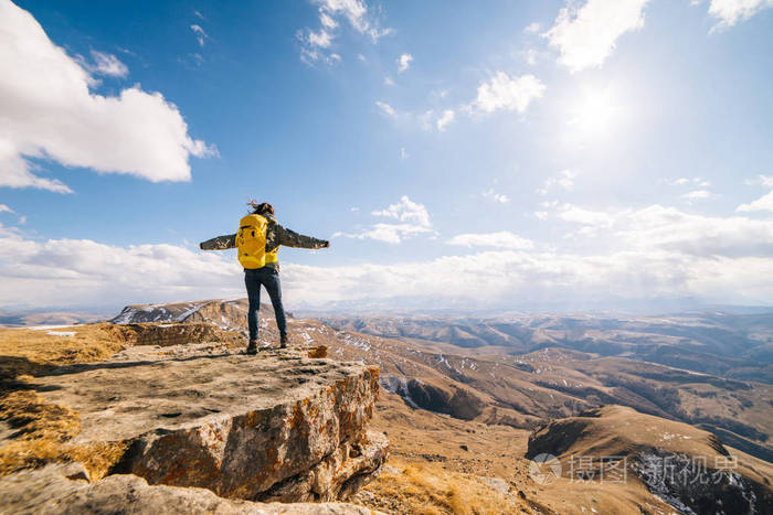 一个活泼的年轻女孩旅行站立在峭壁的边缘并且享受太阳和山干净的空气