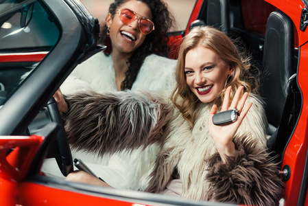 穿着皮大衣拿着车钥匙坐在豪华红车上的快乐时尚多民族女性