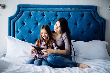 美丽的年轻女子和她迷人的小女儿在家里坐在床上使用数字平板电脑微笑