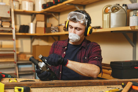 高加索青年男子在格子衬衫, 黑色 t恤, 隔音绝缘耳机, 防护面具工作在木工车间在木桌地方用不同的工具, 锯铁用力量锯
