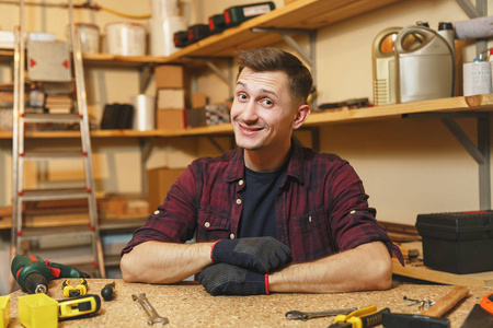 英俊的微笑高加索青年男子在格子衬衫, 黑色 t恤, 在木工车间工作的手套木桌地方与一块木头, 不同的工具
