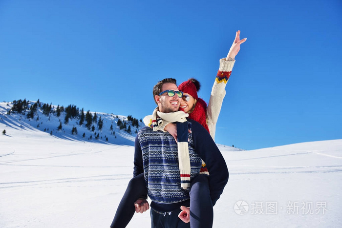 年轻夫妇在雪地上玩得很开心。快乐的男人在山上给他的微笑的女朋友背骑